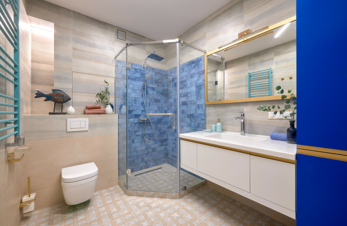 В ванной комнате основным цветом, конечно, выбран голубой. Потолок и часть стен выполнены в этом оттенке. Душевая кабина организована в углу ванной. Трап для слива воды установлен прямо в полу. Зона душа выложена мелкой плиткой сине-голубого цвета. Дверцы кабинки сделаны из прозрачного стекла, тем самым зрительно не уменьшают площадь помещения. Половая плитка уложена по диагонали, за счет этого, ванная комната не вытягивается в длину. Цвет плитки на нижней части стены хорошо подобран по тону с цветом пола. Коврик на полу в коралловом цвете создает ощущение морской растительности. Профильные светильники на потолке задают эффект движения.